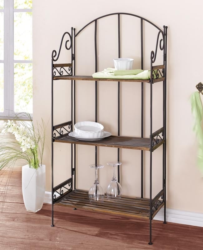 Black metal bakers rack with four shelves holding white dishes, green towels, and glassware in a living room setting