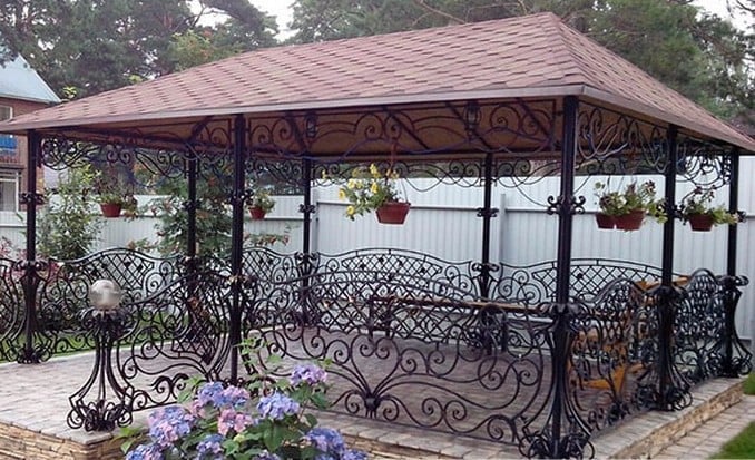 Ornate metal gazebo with copper roof and decorative wrought iron railings in a garden setting with purple flowers