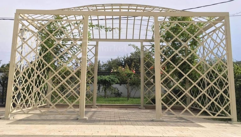 Wooden pergola structure with lattice panels on sides, paved ground, trees and greenery in background
