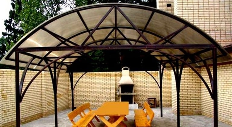 Metal pergola structure with arched roof over a courtyard patio area, featuring wooden picnic tables and an outdoor fireplace against a brick wall