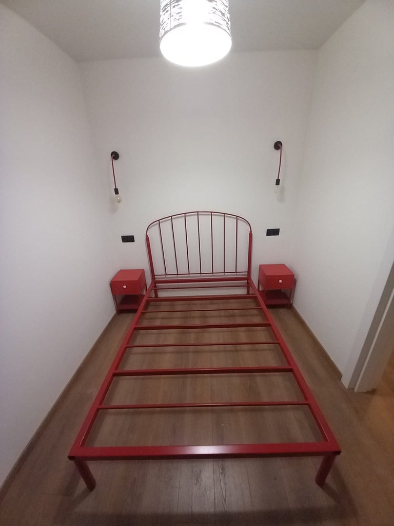 Red metal bed frame with footboard in empty bedroom with white walls, ceiling light, and wooden floor