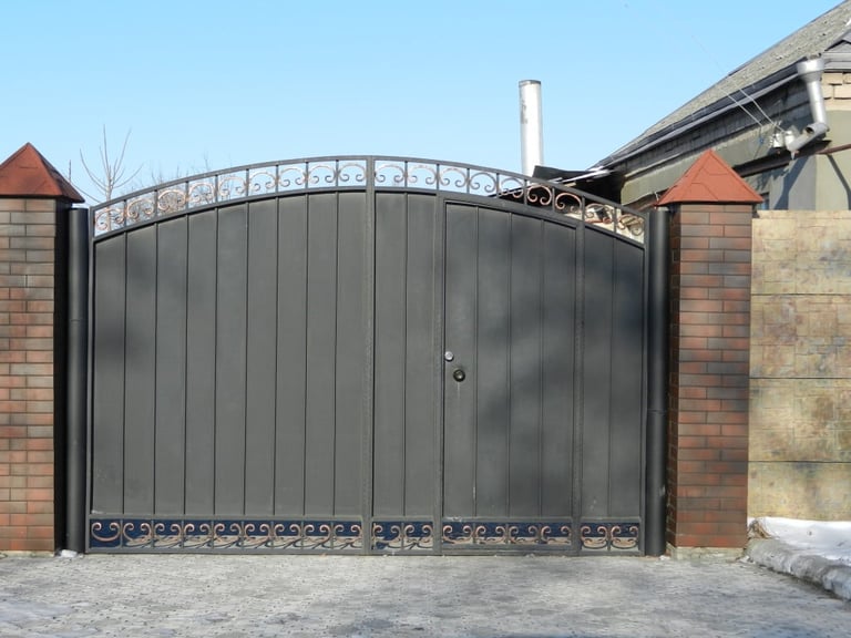 Large ornamental black metal driveway gate with curved top between brick pillars and stone walls