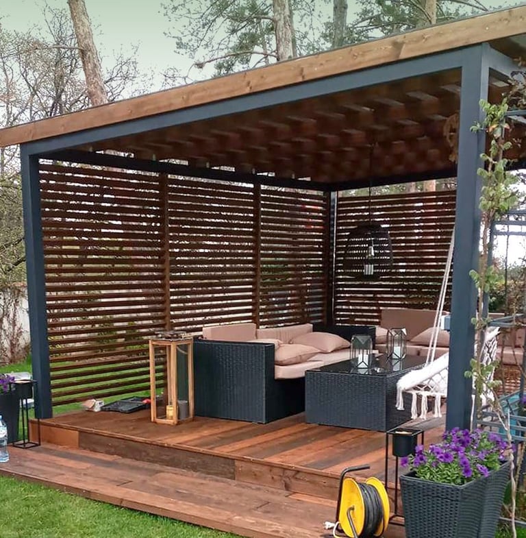 Modern backyard pergola with wooden deck, black lattice panels, wicker seating with pink cushions, and purple flowers in planters