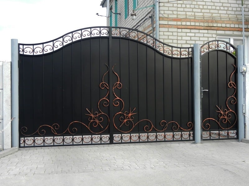 Black ornamental metal driveway gate with decorative gold flourishes and matching pedestrian gate on brick building facade