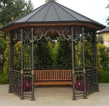 Ornate metal hexagonal gazebo with a conical roof and wooden bench interior, set in a garden